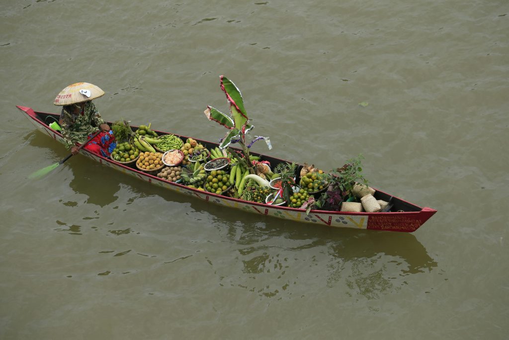 Une femme sur une barque flottant sur un lac et qui transporte une variété de légumes et fruits 